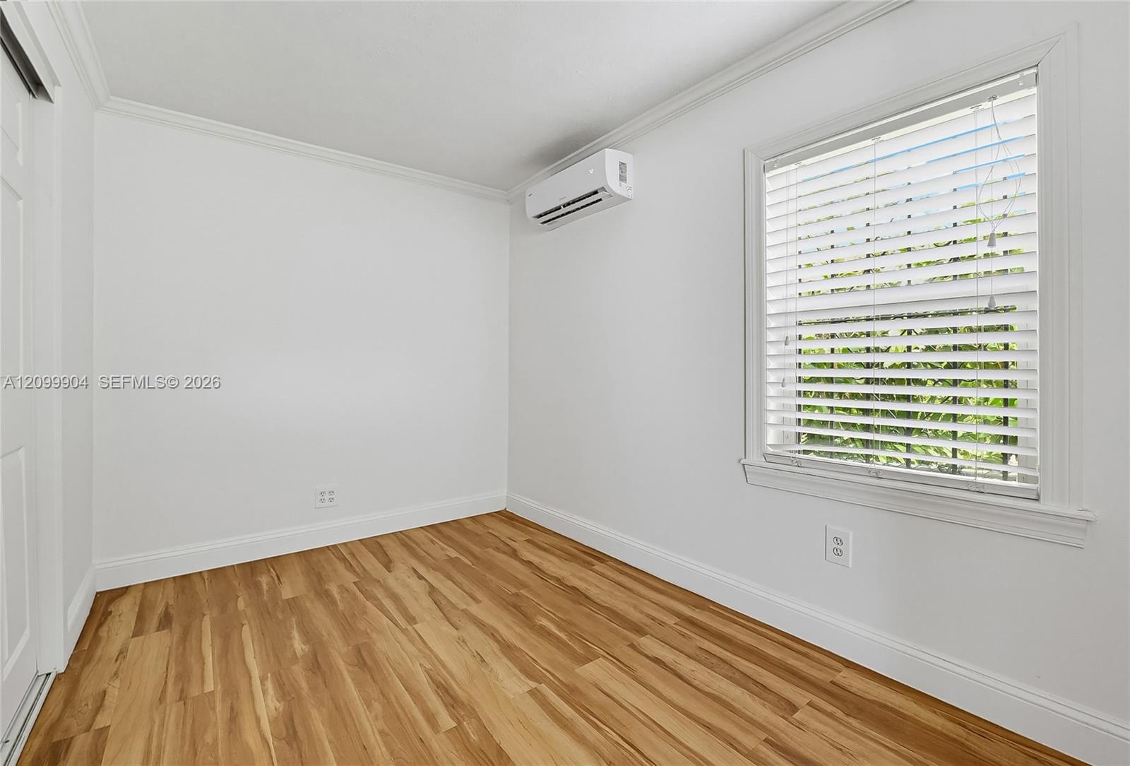 Wood-finish flooring, crown molding, wall-mounted ductless air conditioning unit, and a window with horizontal blinds