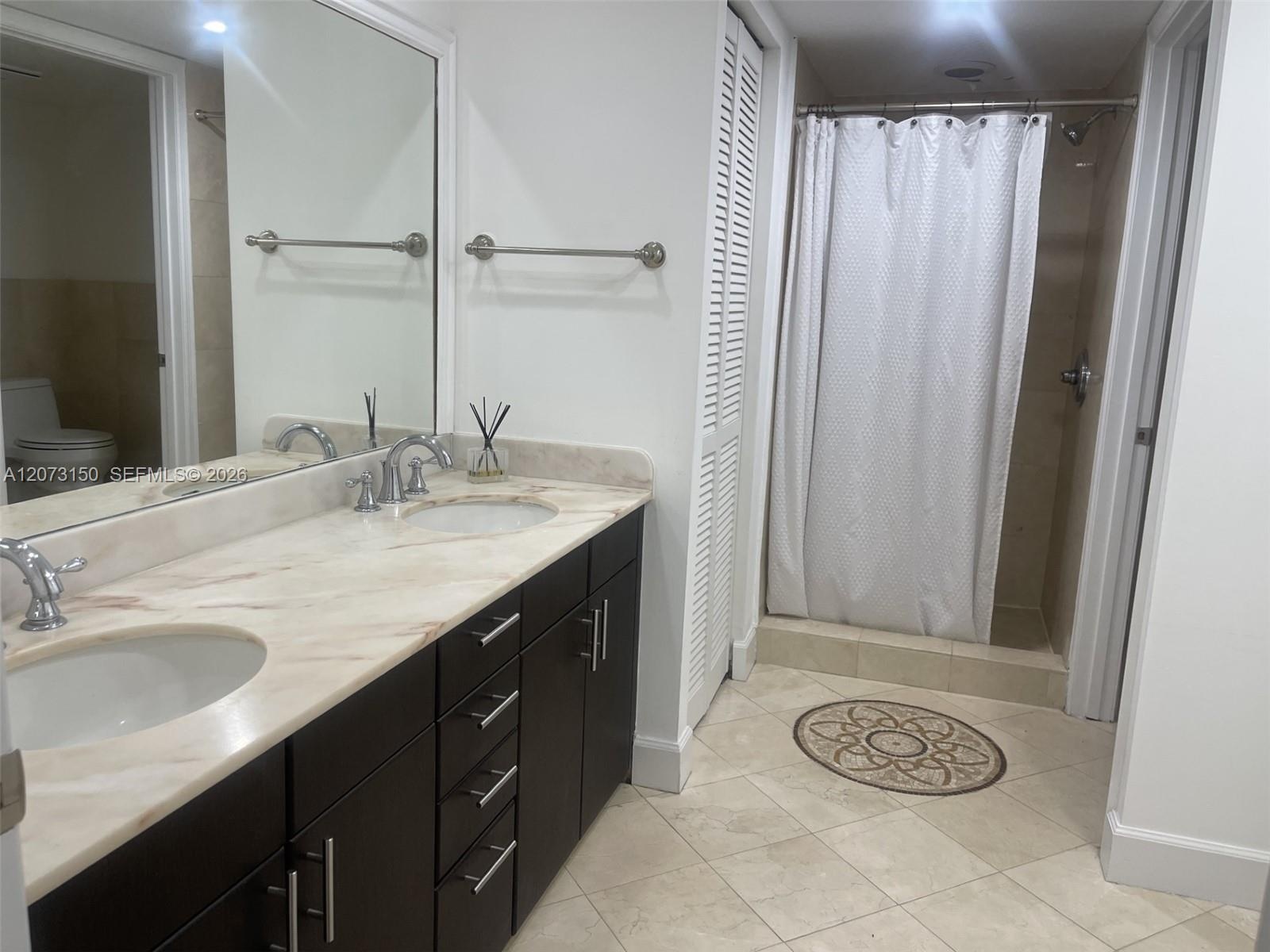 Dual-sink vanity featuring stone countertop, dark wood-finish cabinetry, polished metal fixtures, and walk-in shower with tile surround