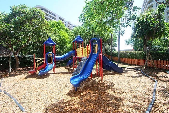 Playground area featuring multi-level play structures, curved slides, stone-textured climbing element, wood chip ground cover, and mature trees