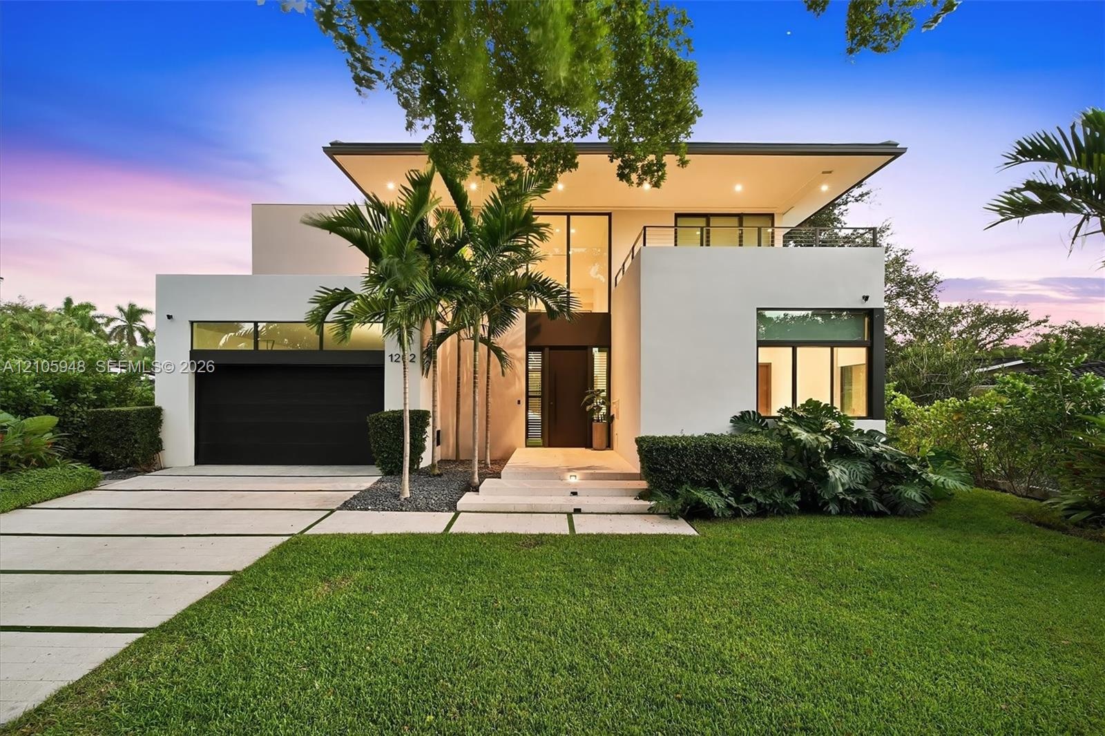 Modern architectural exterior featuring a flat roofline, oversized glass entry, second-level balcony with metal railing, paver driveway, and recessed 