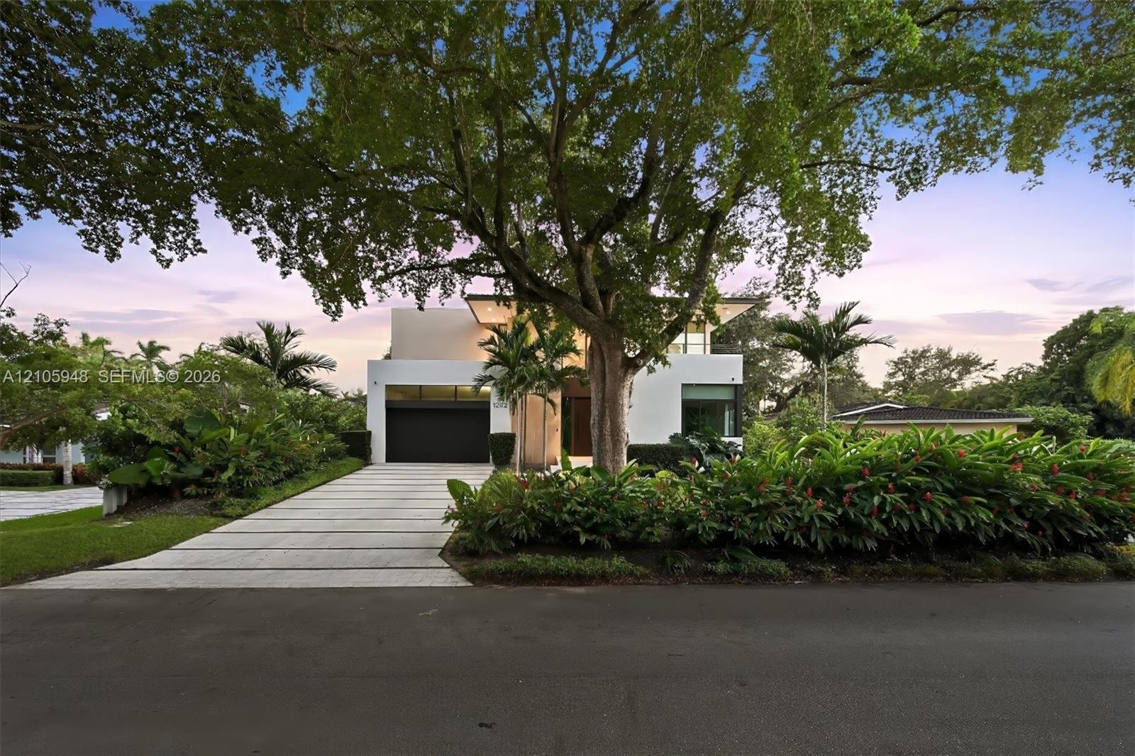 Modern architectural facade featuring a smooth stucco exterior, linear concrete paver driveway, dark-finish multi-car garage door, and lush tropical l