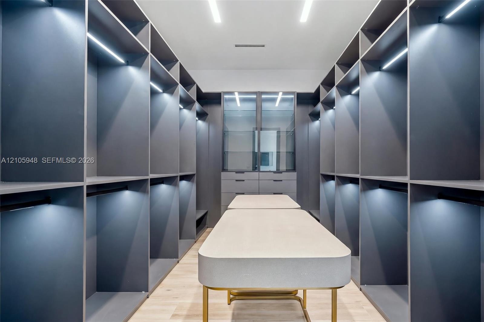 Custom walk-in wardrobe featuring floor-to-ceiling matte cabinetry, integrated LED shelving, glass-fronted display cases, a central island with a ston