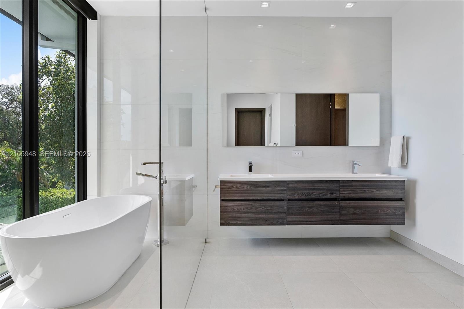 Freestanding oval soaking tub, wall-mounted dark wood-finish dual vanity with integrated sinks, large-format tile flooring, floor-to-ceiling glass-enc