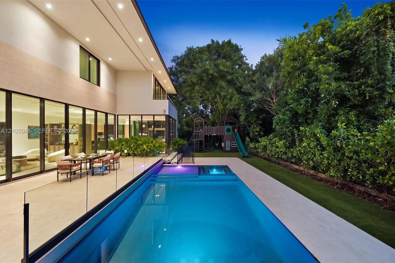 Rear exterior featuring floor-to-ceiling glass wall panels, stone-finish patio, rectangular swimming pool with integrated spa, glass railing, and mani