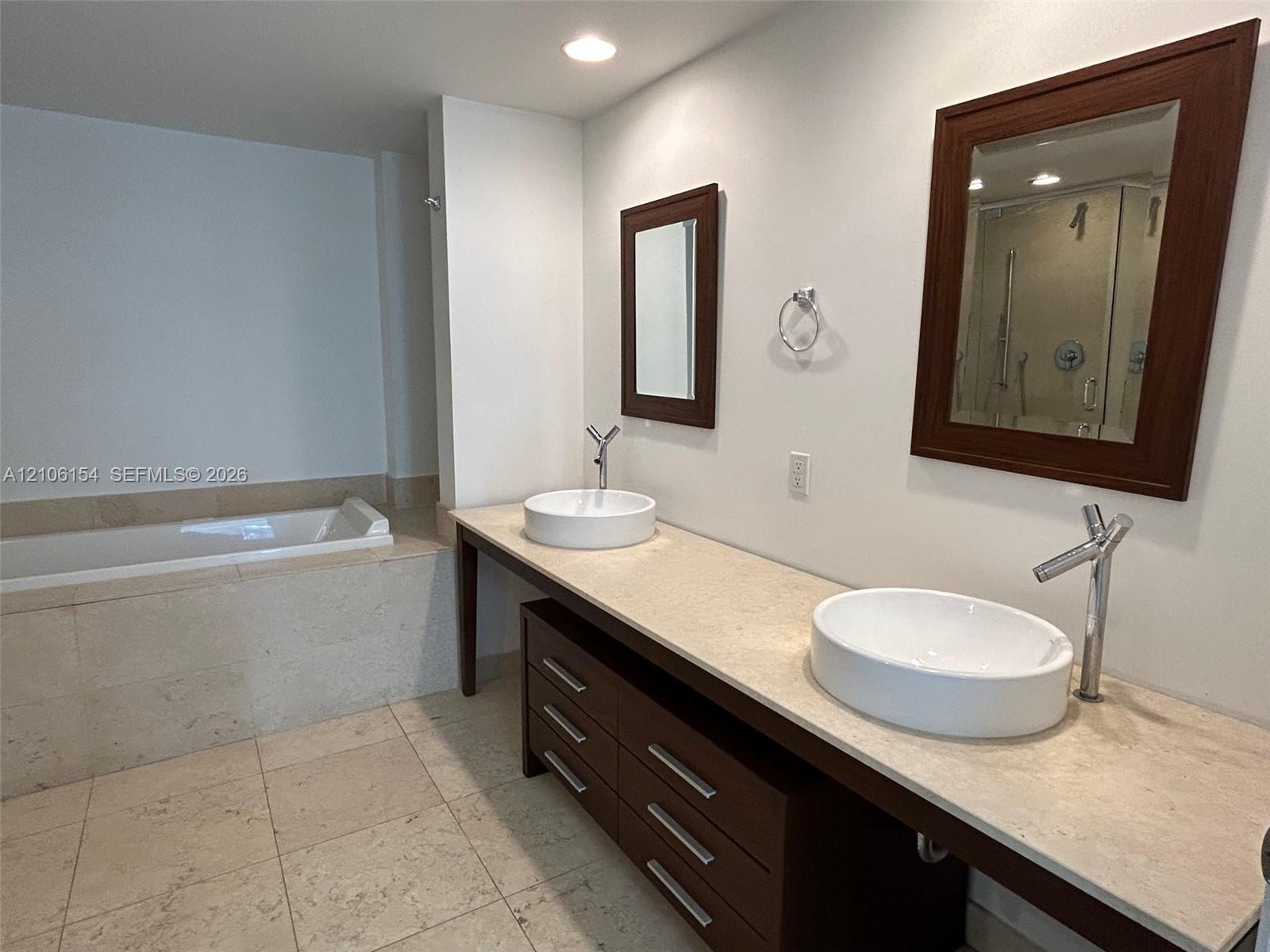 Double vanity featuring a light-toned stone countertop, dual vessel sinks, dark wood-finish cabinetry, and a deep soaking tub with a stone tile surrou