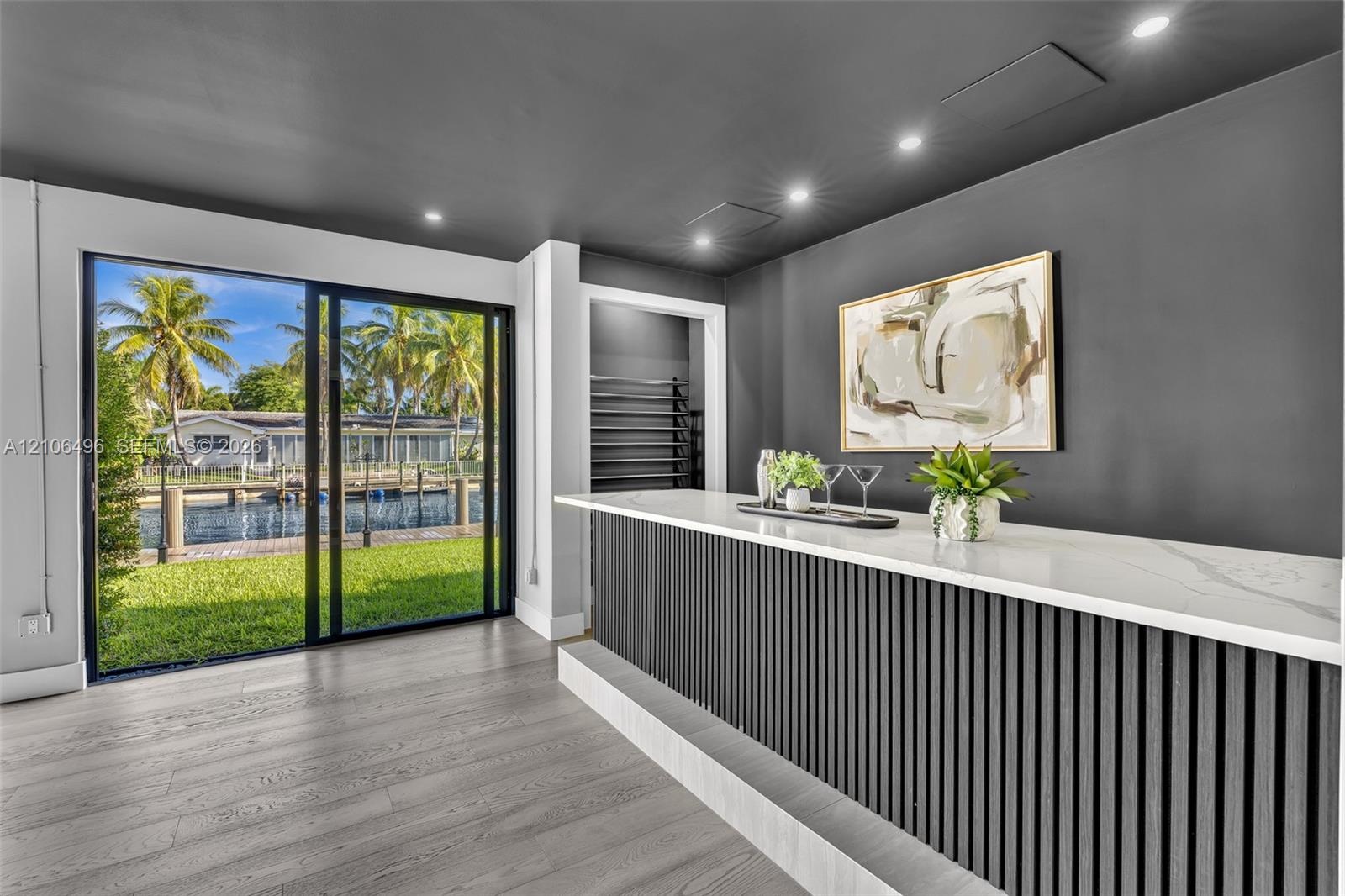 Built-in bar featuring a stone-surface countertop, slatted wood-finish base, recessed ceiling lighting, and sliding glass doors with water and landsca