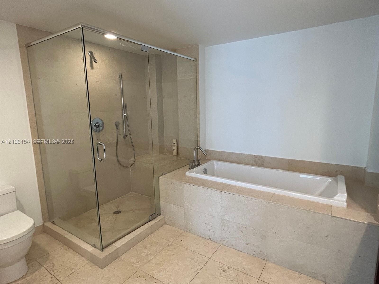 Walk-in glass-enclosed shower with integrated seating, drop-in soaking tub with stone tile surround, and neutral stone tile flooring
