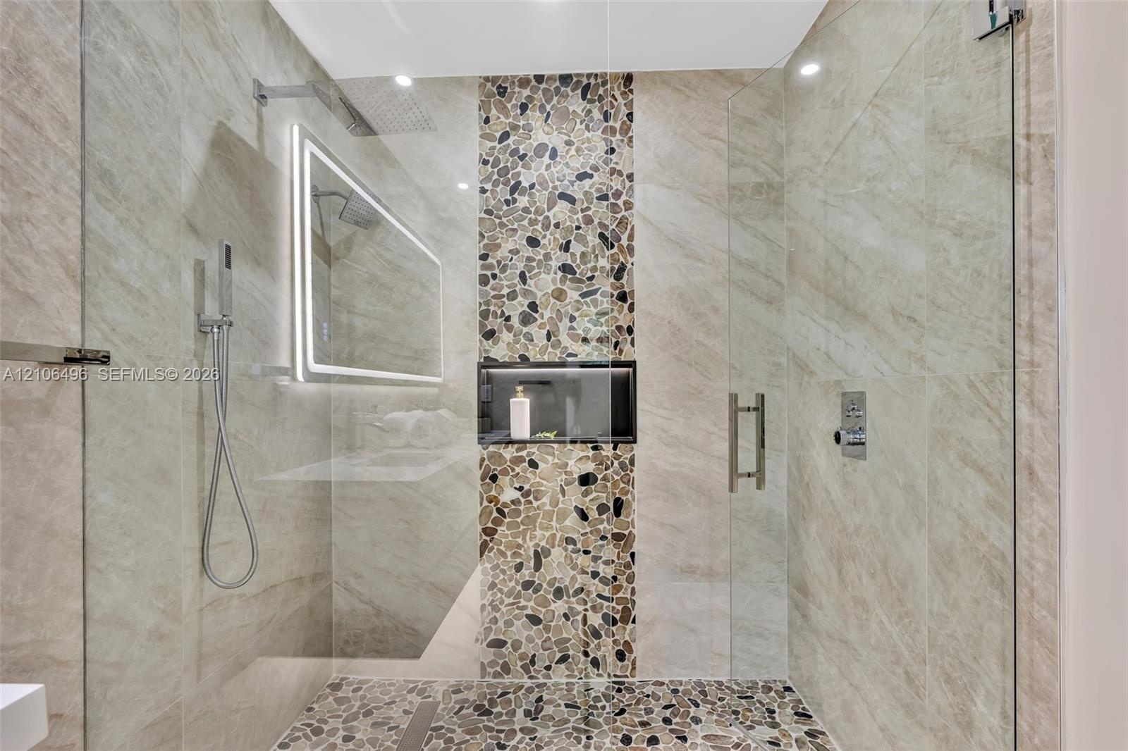 Walk-in shower featuring large-format neutral wall tile, a vertical pebble-stone accent column, pebble-tile flooring, a recessed storage niche, and an
