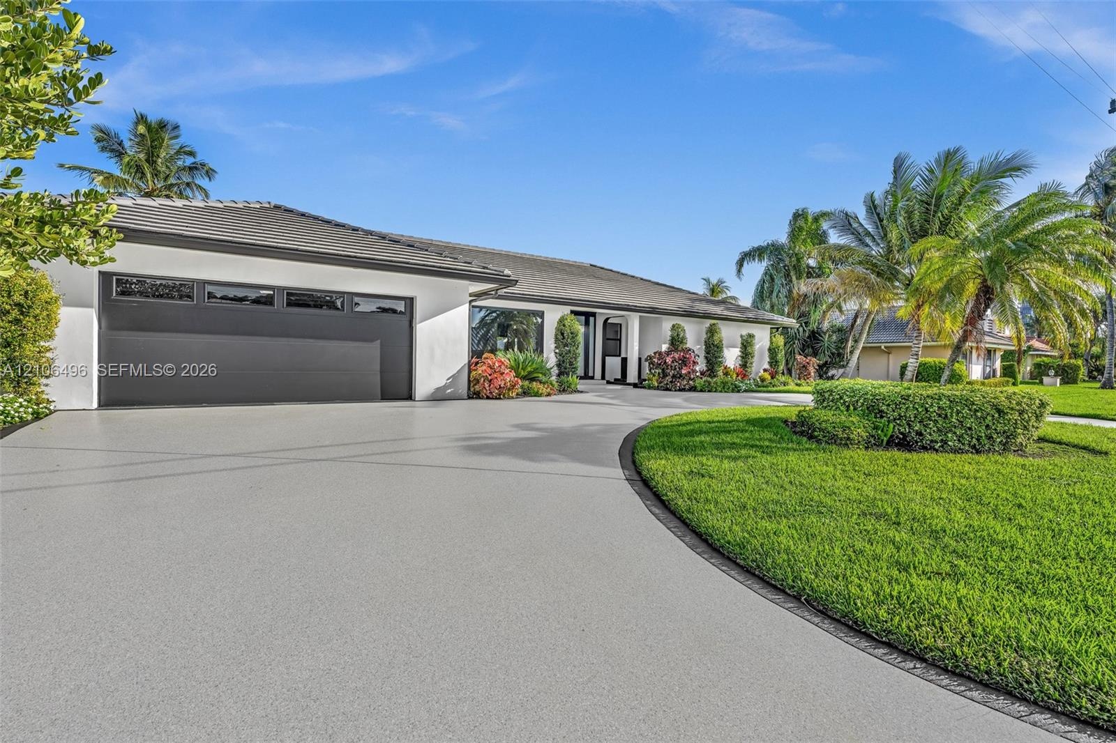 Single-story exterior featuring white stucco siding, a multi-car garage with overhead glass-paneled door, a curved concrete driveway, and mature lands