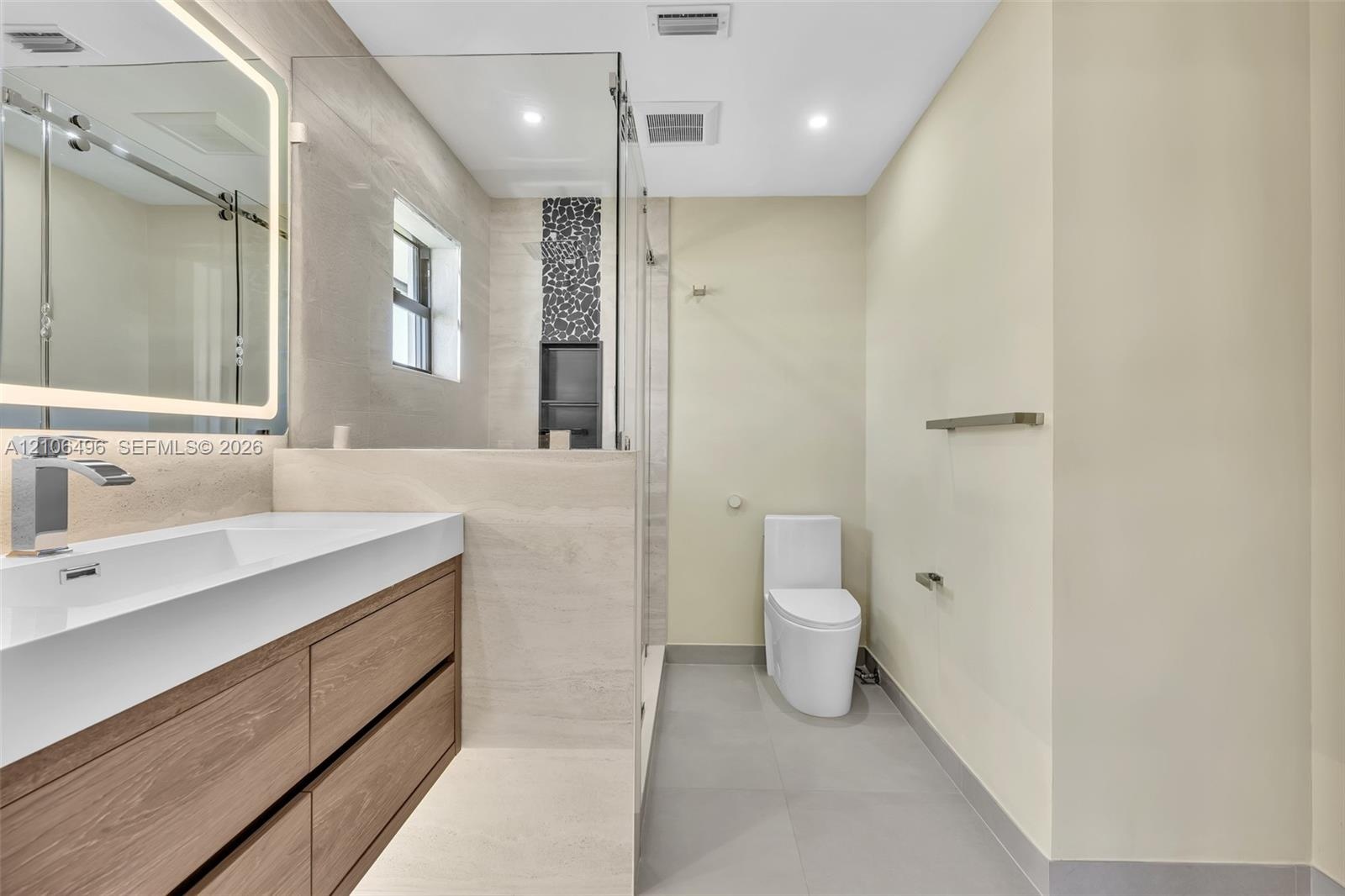 Floating wood-finish vanity with an integrated rectangular sink, backlit vanity mirror, glass-enclosed shower with decorative stone-look inlay, and la
