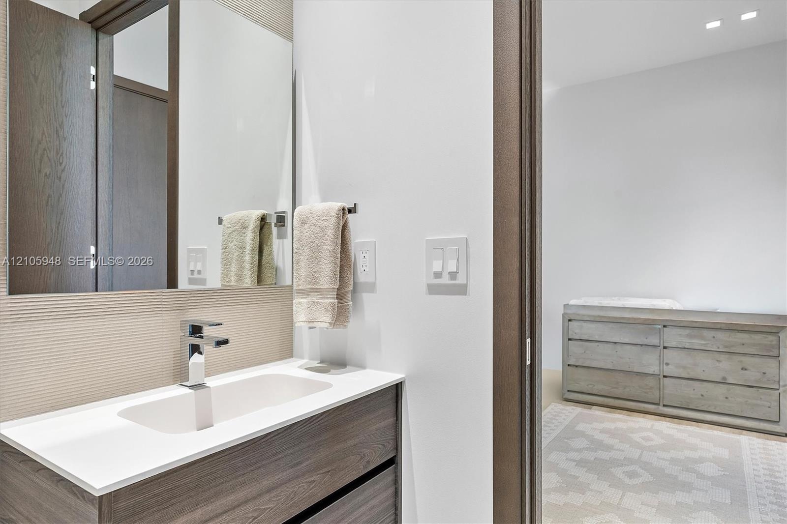 Wall-mounted mirror above a vanity featuring a wood-finish cabinet, white countertop, integrated rectangular sink, and polished chrome faucet