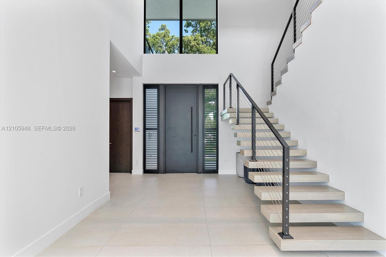 Grand foyer featuring a double-height ceiling, floor-to-ceiling windows, modern floating staircase with cable railing, large-format tile flooring, and
