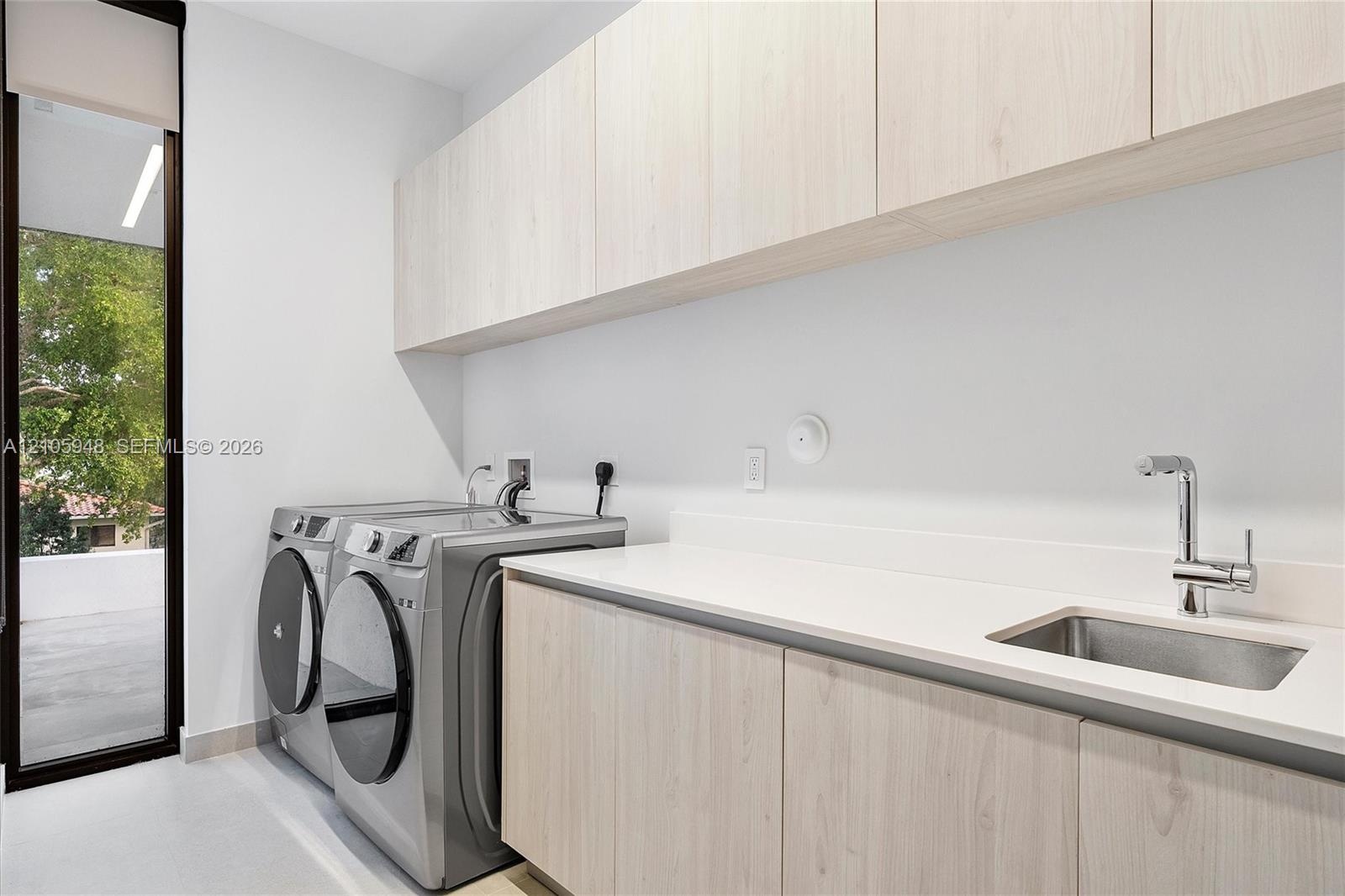 Laundry space featuring light-toned cabinetry, solid-surface countertops, an integrated stainless steel sink with a high-arch faucet, and full-height 