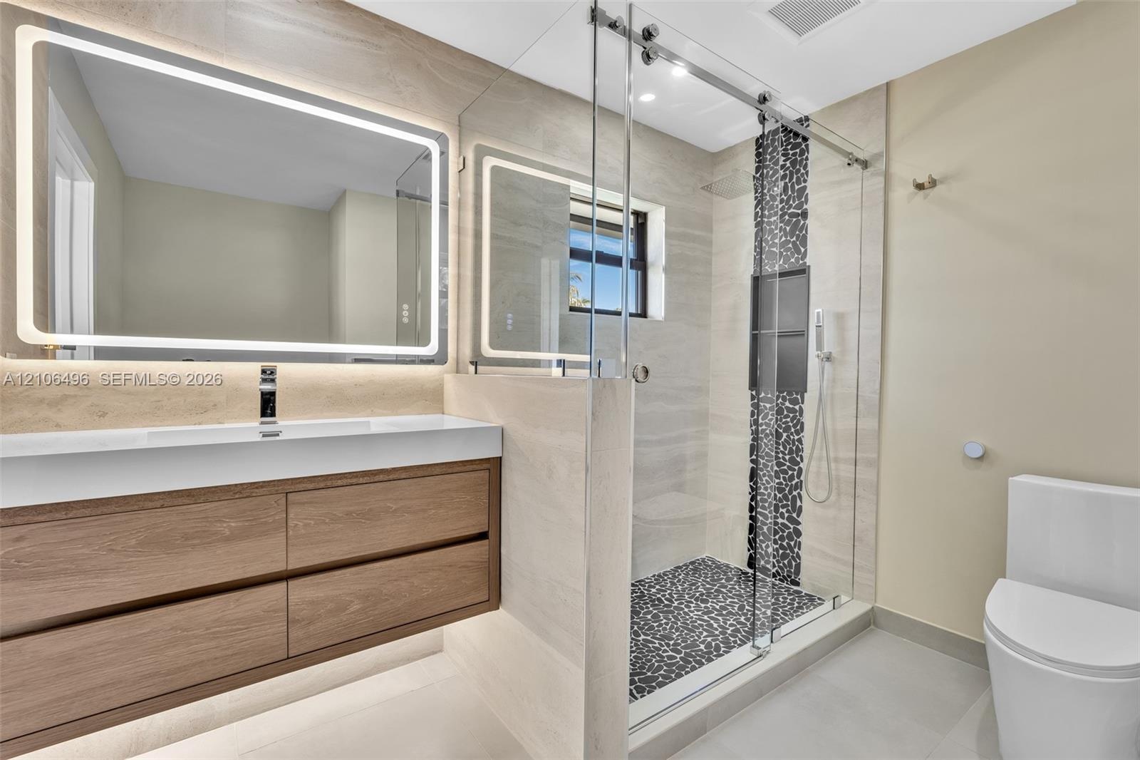 Floating wood-finish vanity with an integrated countertop, large LED-backlit mirror, and walk-in shower featuring glass enclosure, pebble stone floori