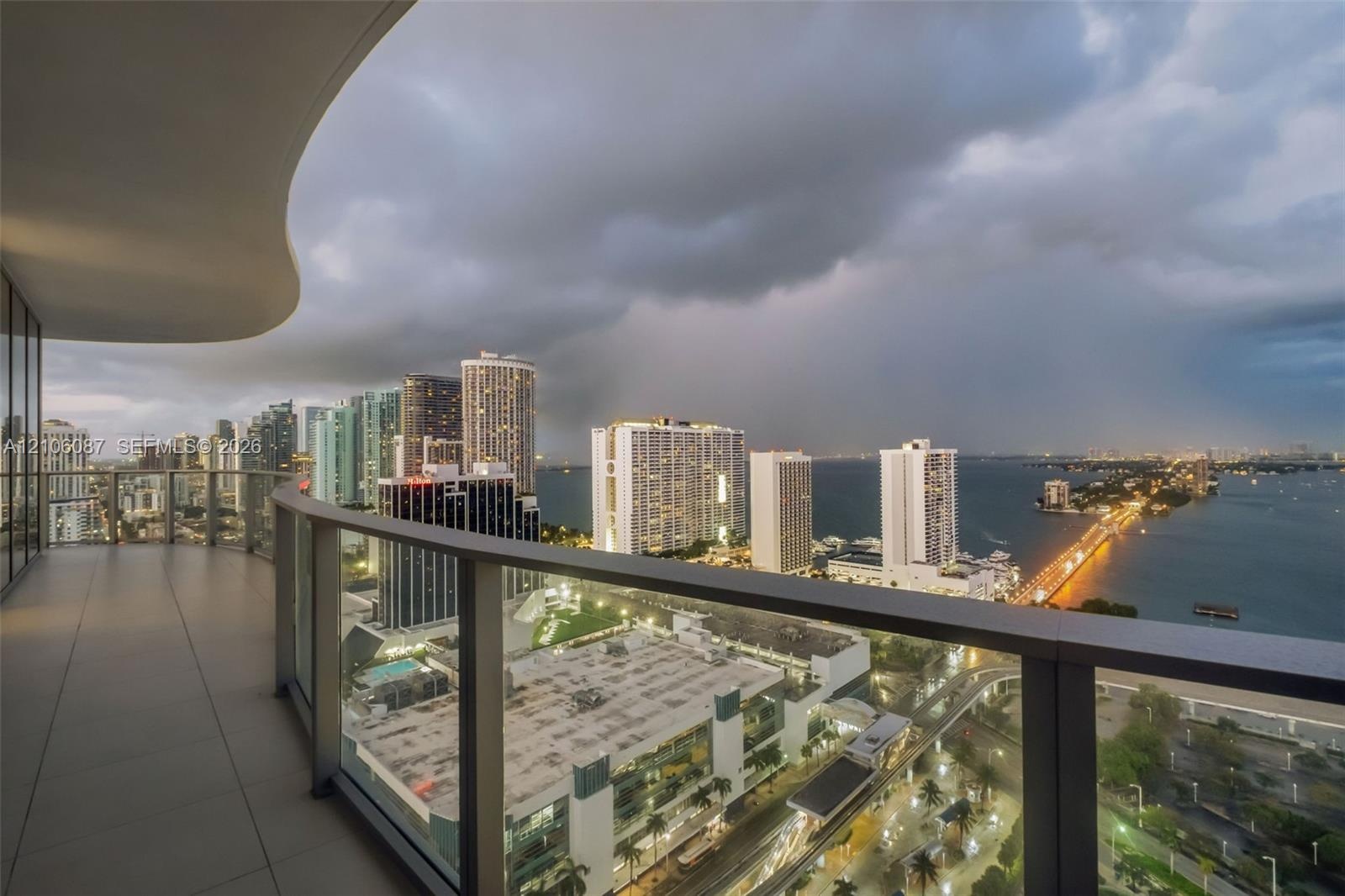 Curved balcony with large-format tile flooring and a glass-paneled railing overlooking an urban skyline and waterfront