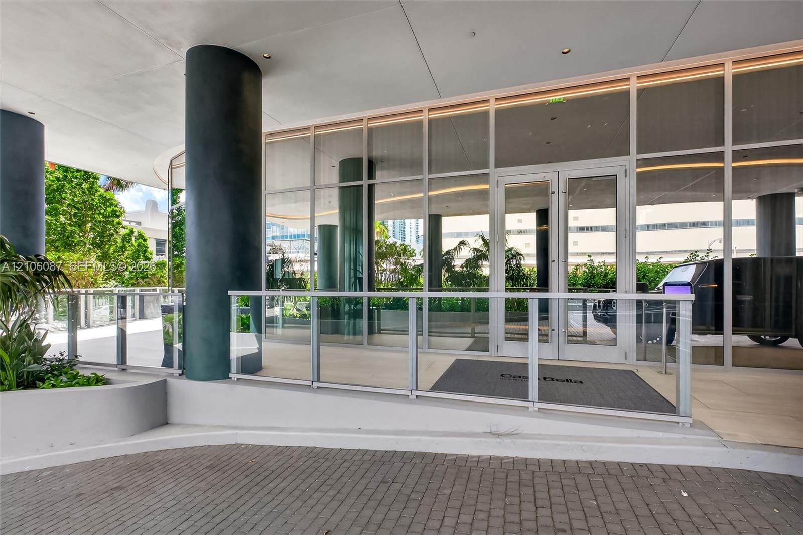 Covered porte-cochère entry featuring floor-to-ceiling glass paneling, cylindrical structural columns, stone-paver driveway, and a glass-paneled raili