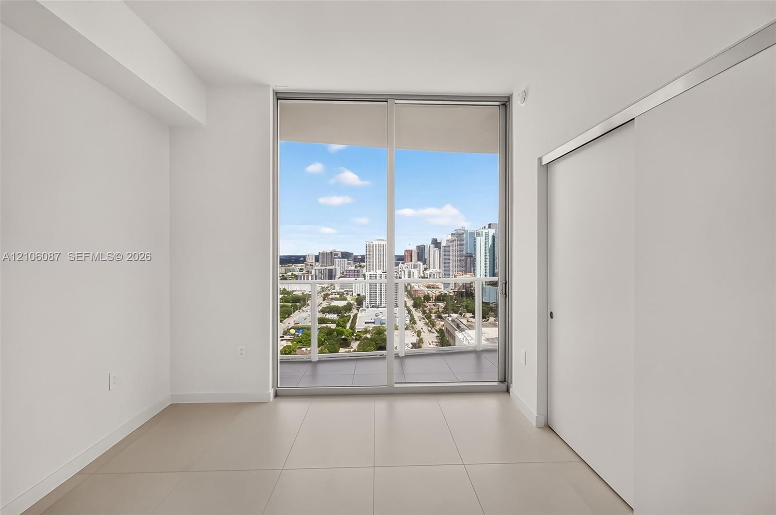 Floor-to-ceiling sliding glass doors, private balcony, neutral large-format tile flooring, and urban skyline backdrop