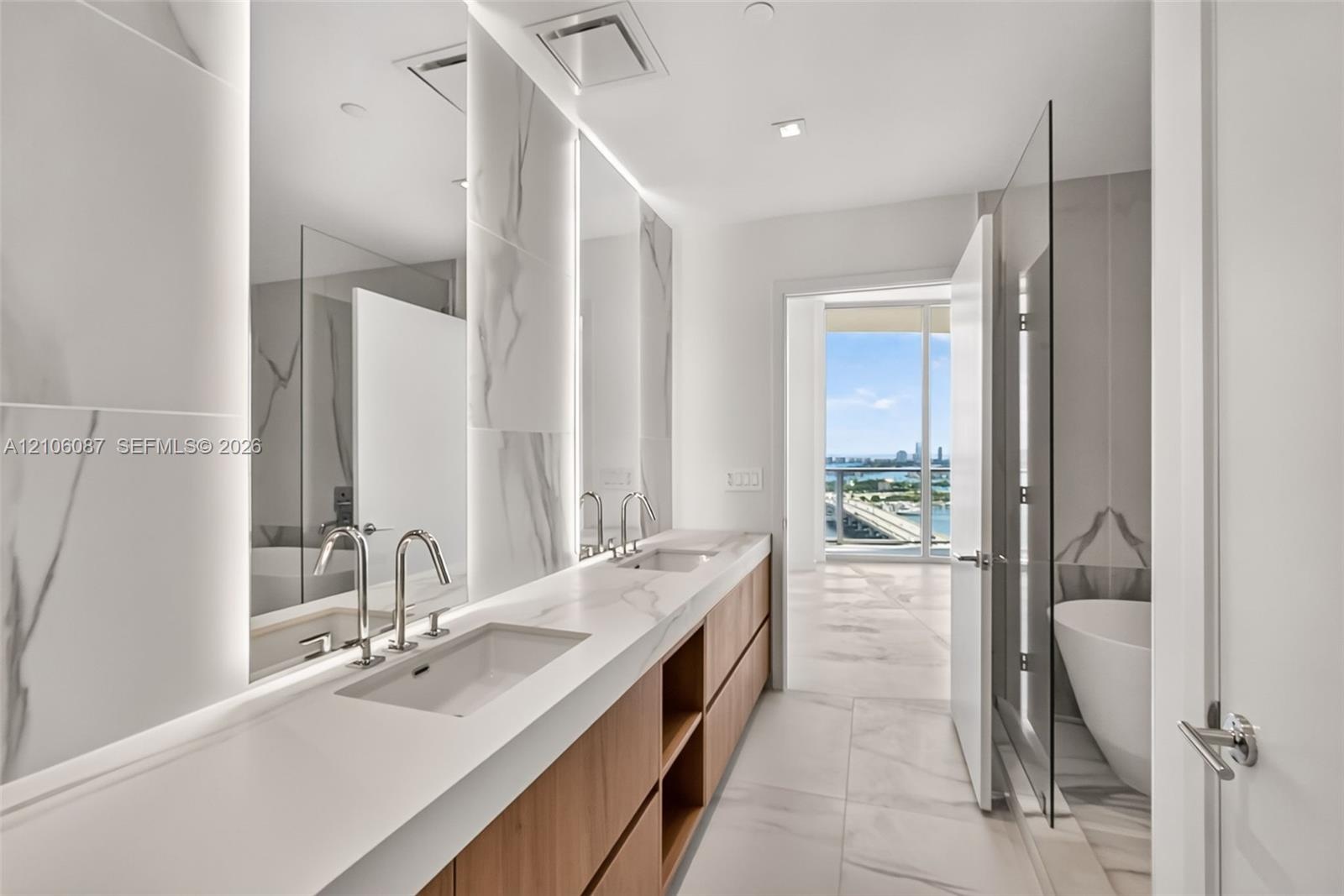 Double vanity featuring light-toned wood cabinetry, large-format tile flooring, integrated wall-mounted mirrors with backlighting, and a glass-enclose