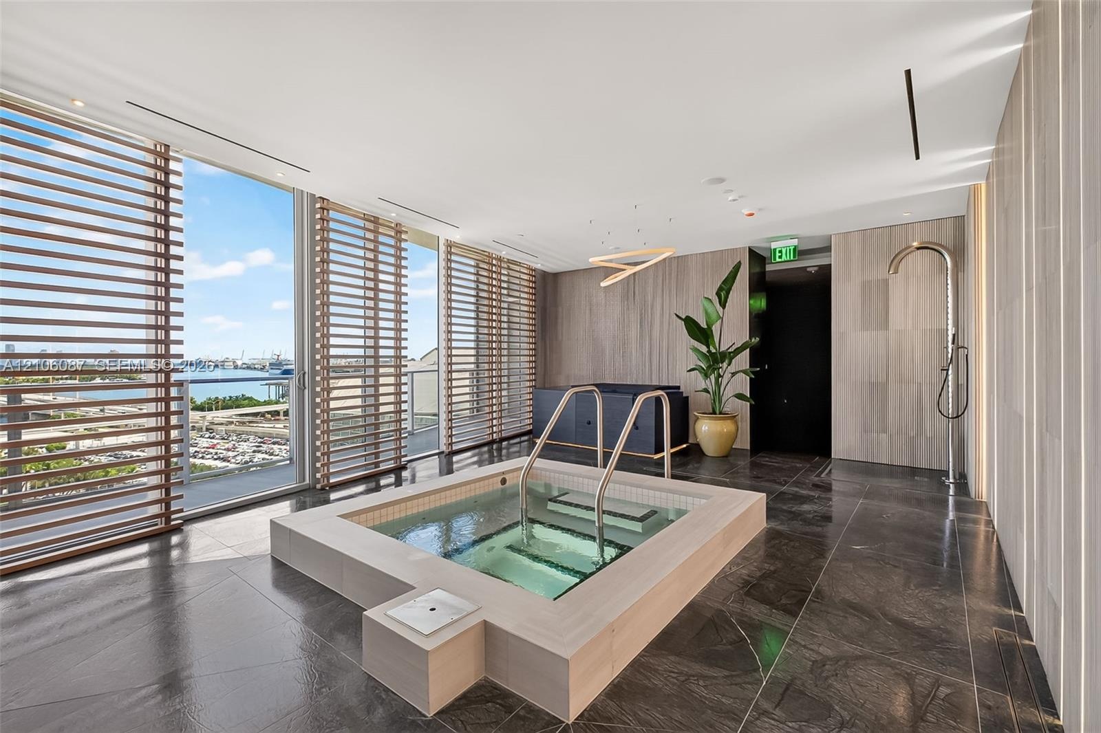 Recessed soaking tub featuring a stone-tile surround, floor-to-ceiling windows with wood-slat screening, large-format stone floor tiles, and a wall-mo
