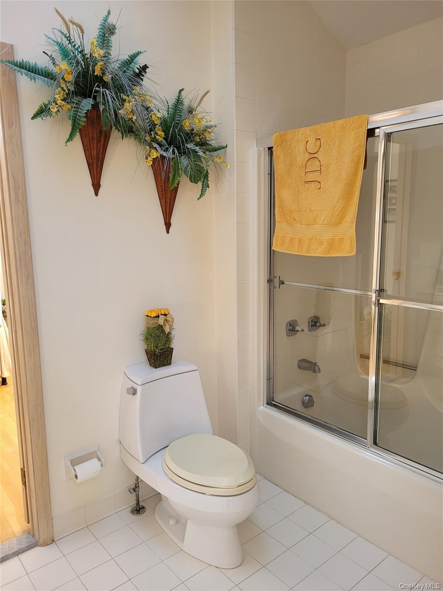 White tile flooring with a matching toilet, a built-in shower-tub combination, and a glass shower enclosure