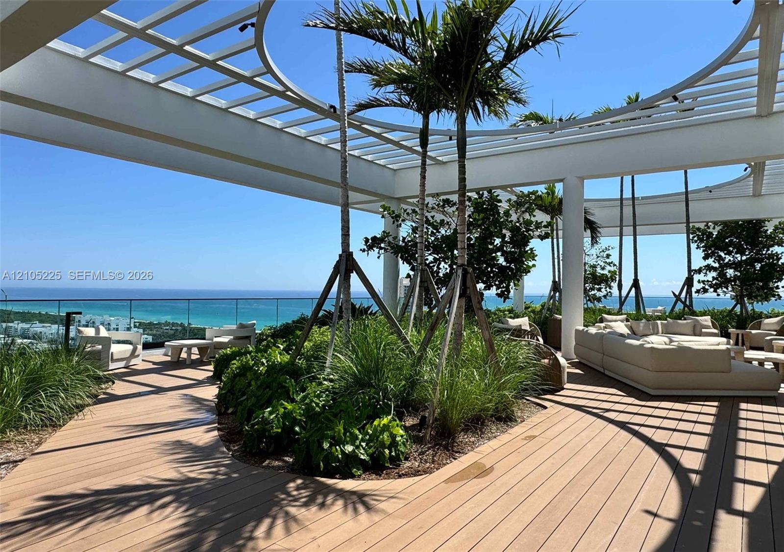 Expansive outdoor terrace featuring wood-finish decking, a white slatted pergola, lush landscaping, and a clear glass railing