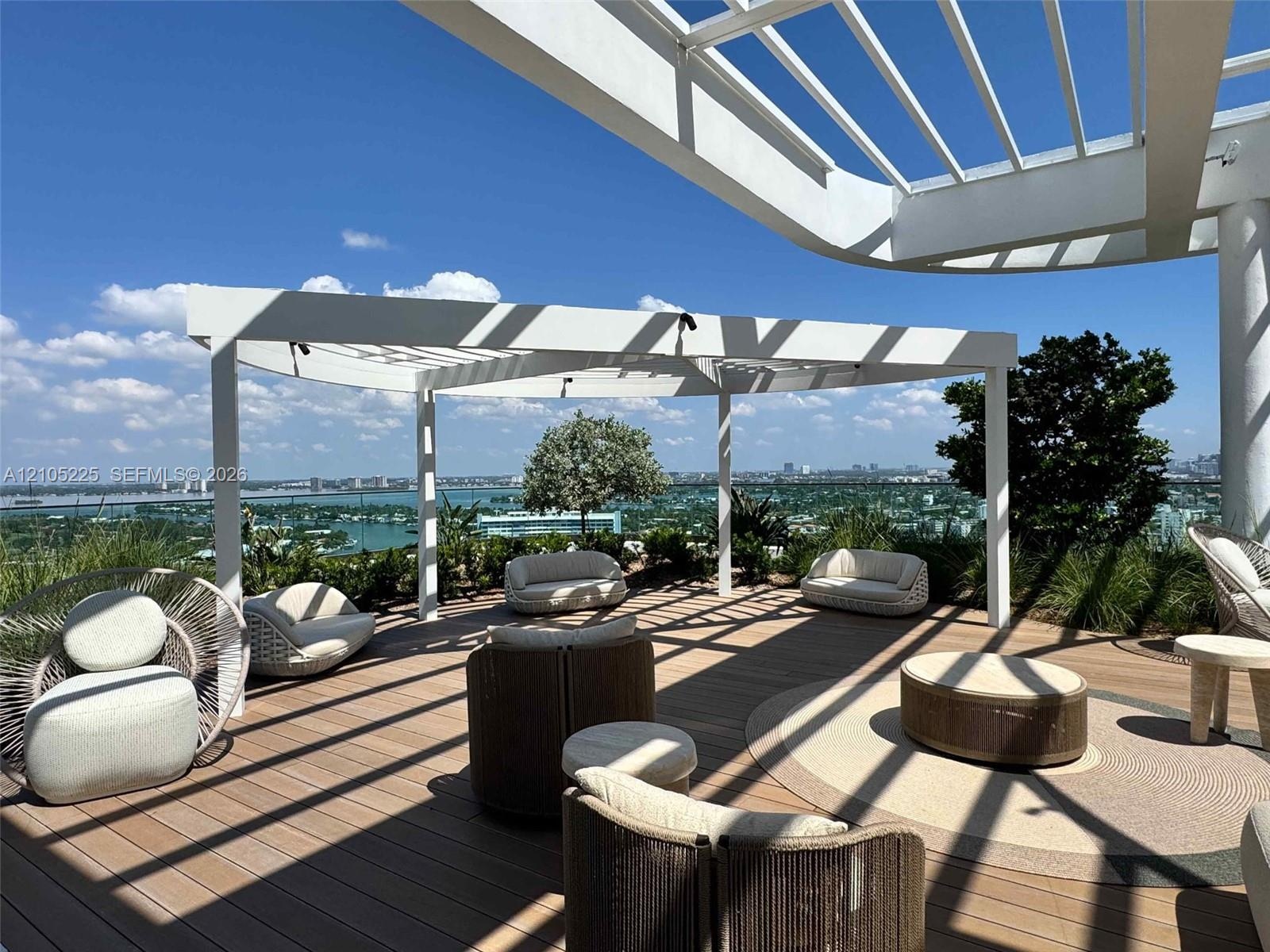 Expansive rooftop terrace with wood-finish decking, white pergola structure, integrated landscaping, and distant city and water scenery