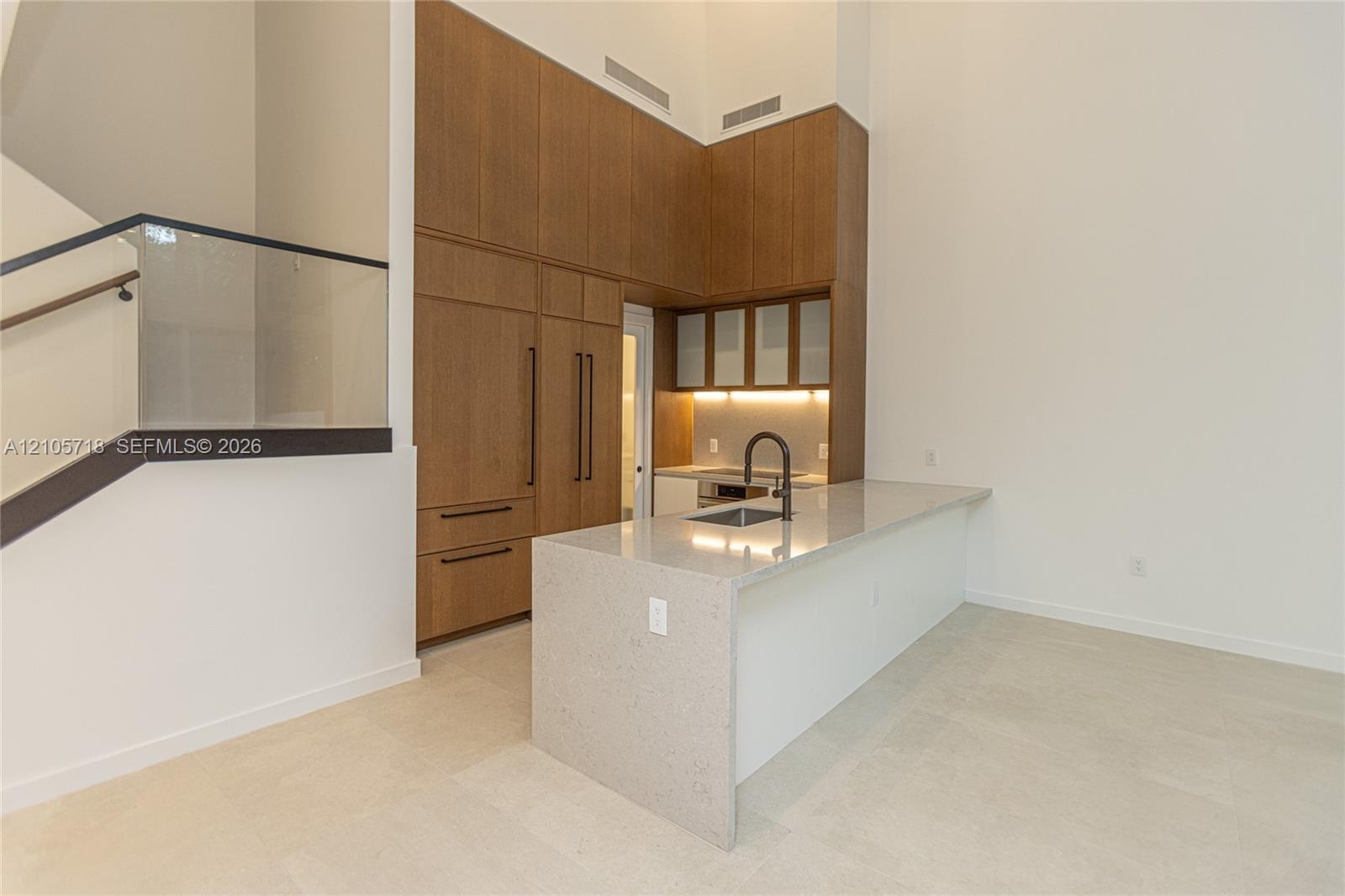 Kitchen featuring full-height wood-finish cabinetry, waterfall-edge stone countertop, undermount sink with matte black faucet, and glass staircase rai