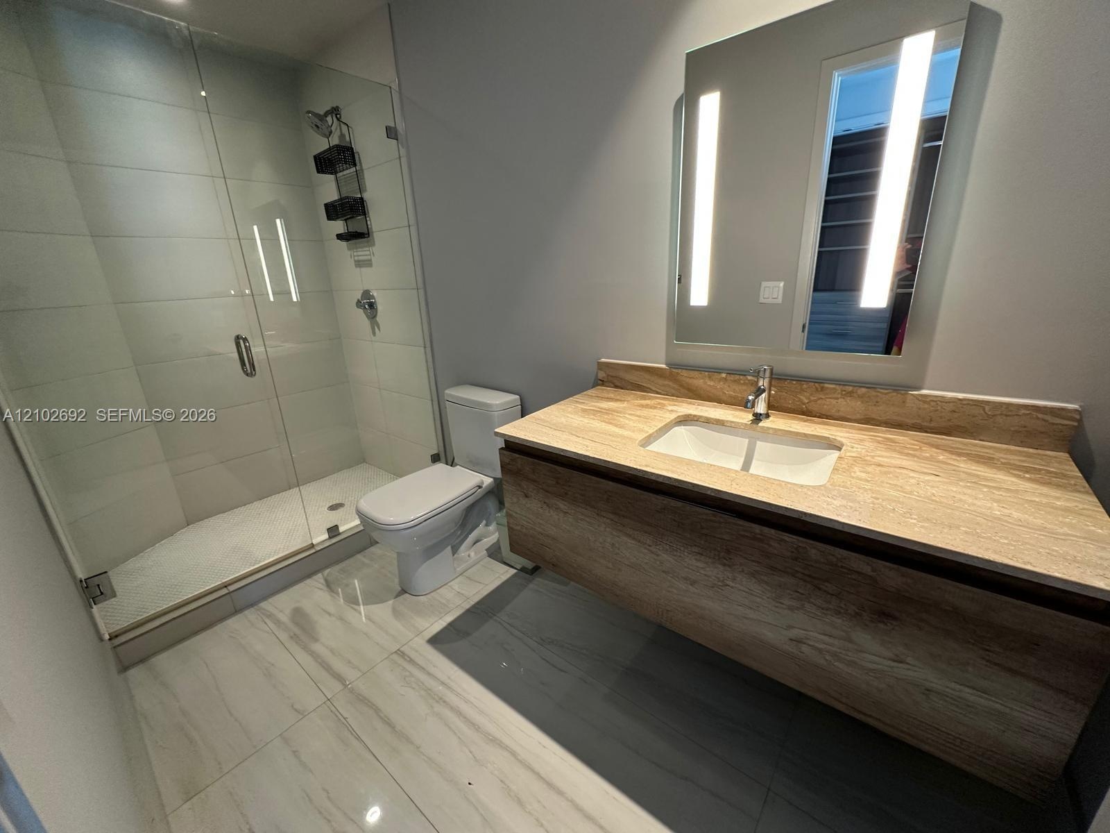 Floating wood-finish vanity with an integrated countertop sink, wall-mounted mirror with integrated lighting, glass-enclosed shower, and large-format 