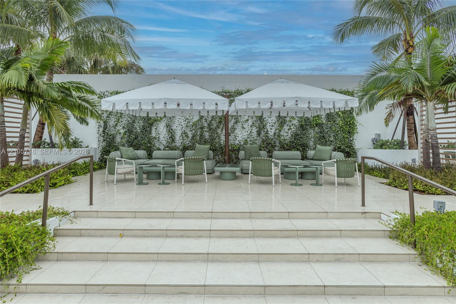 Outdoor terrace featuring oversized light-colored tiling, a tiered staircase with metal handrails, a climbing vine wall, and perimeter tropical landsc