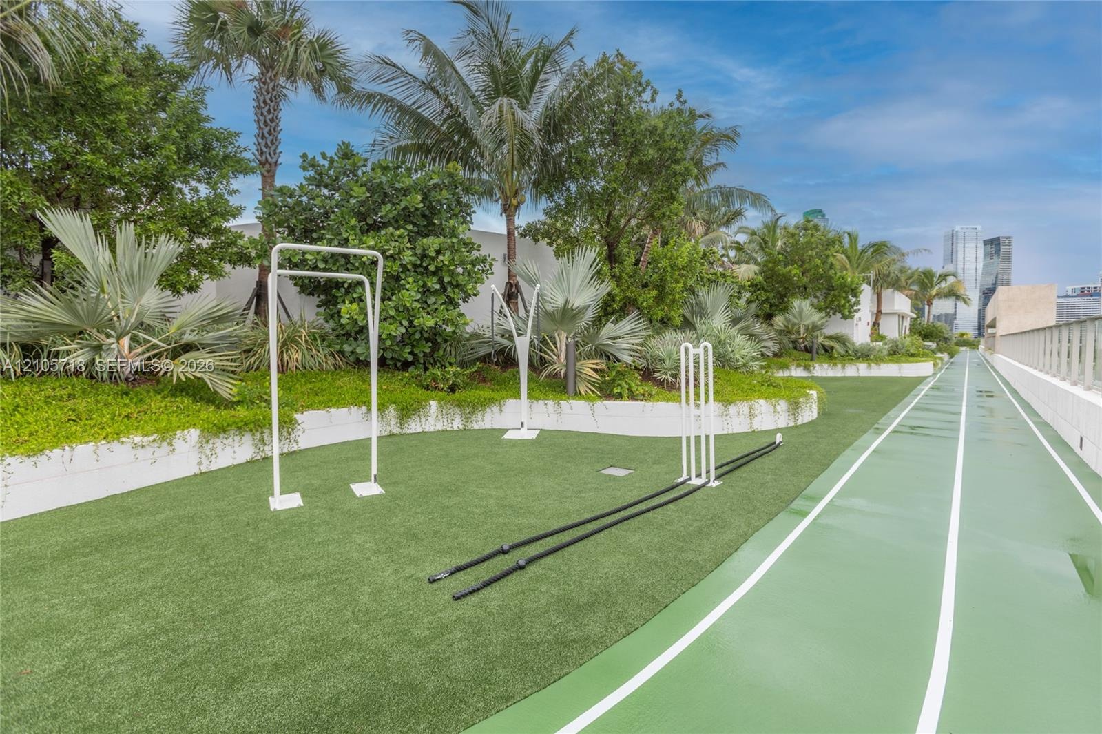 Outdoor fitness area featuring a synthetic running track, multi-use exercise frames, manicured landscaping with mature palms, and tiered retaining wal