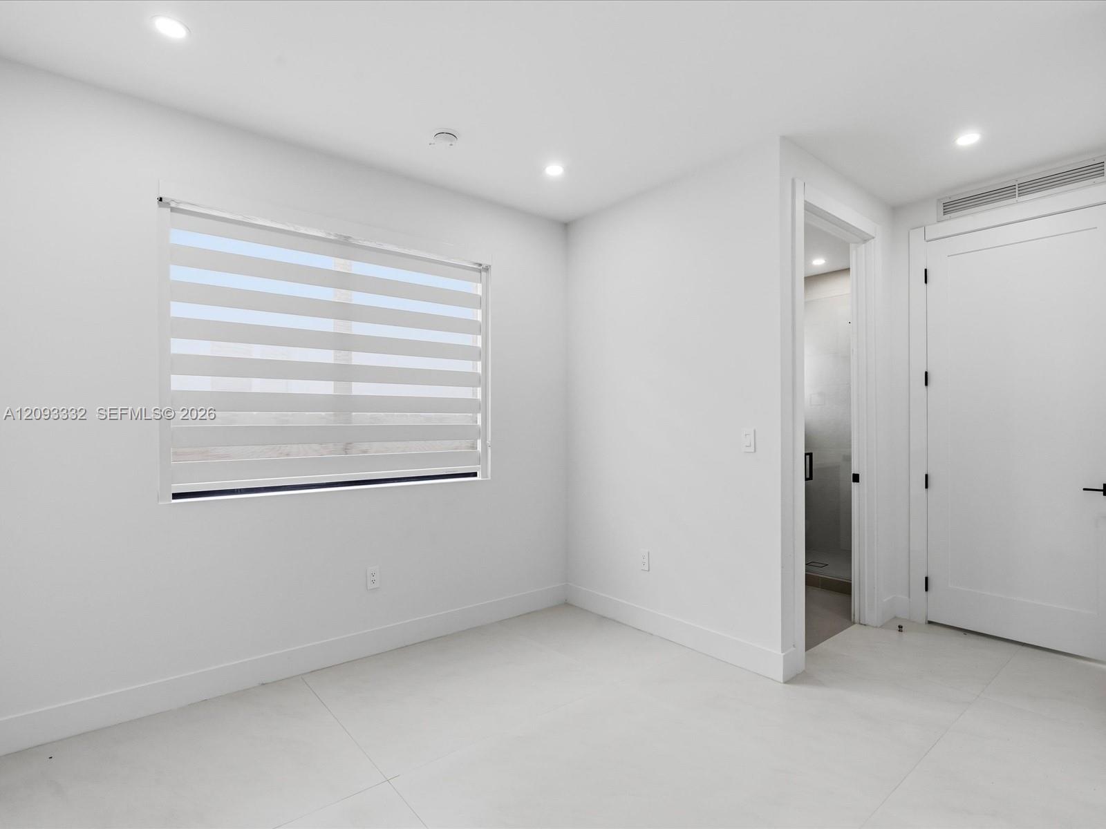 Neutral-toned interior featuring large-format tile flooring, recessed ceiling lighting, a single window with horizontal privacy blinds, and a solid-co