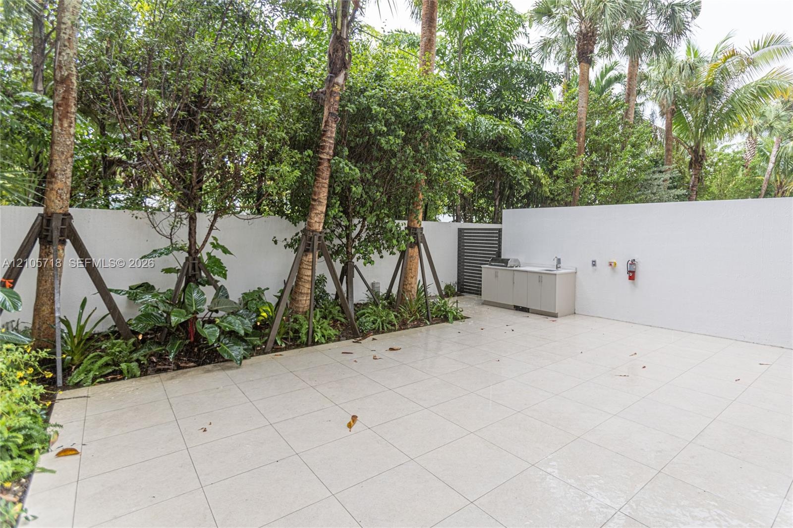 Private outdoor patio featuring light-toned tile flooring, built-in cabinetry with a sink, stainless steel grill, solid privacy walls, and mature peri