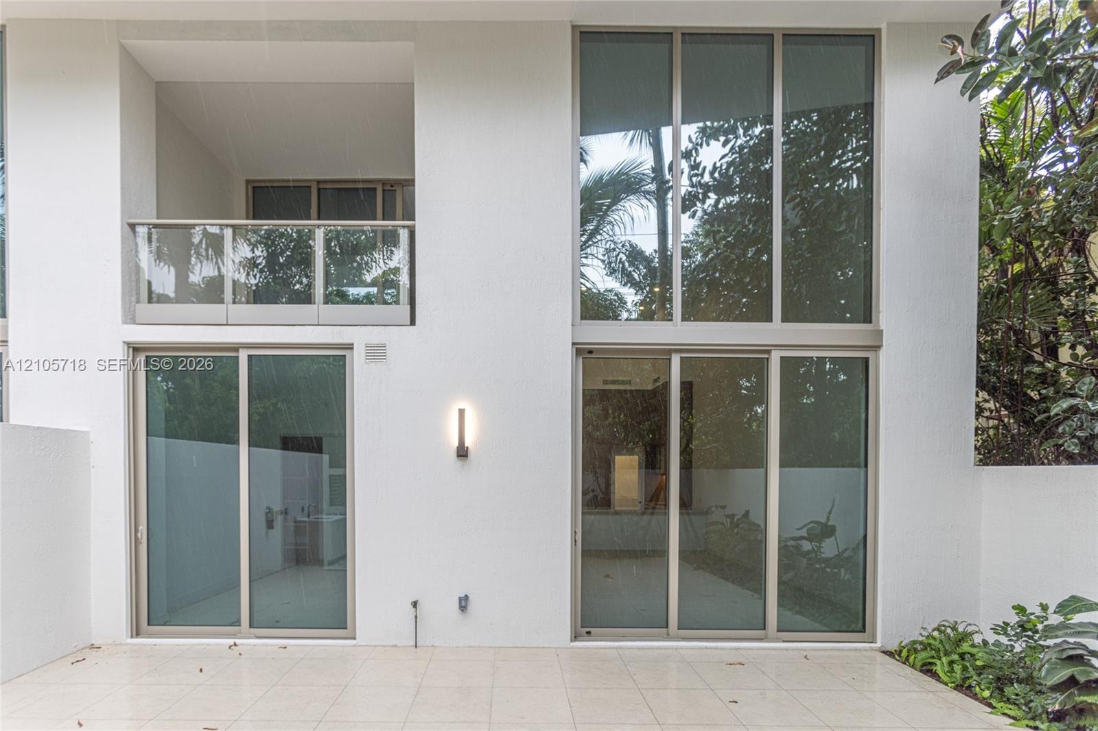Exterior elevation featuring expansive two-story floor-to-ceiling windows, a covered balcony with a glass railing, large-format tile patio flooring, a