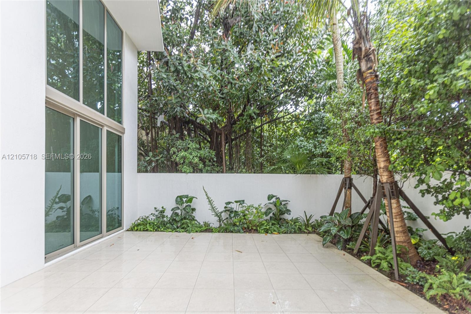 Patio featuring oversized light-toned tile flooring, floor-to-ceiling windows, white perimeter wall, and established perimeter landscaping