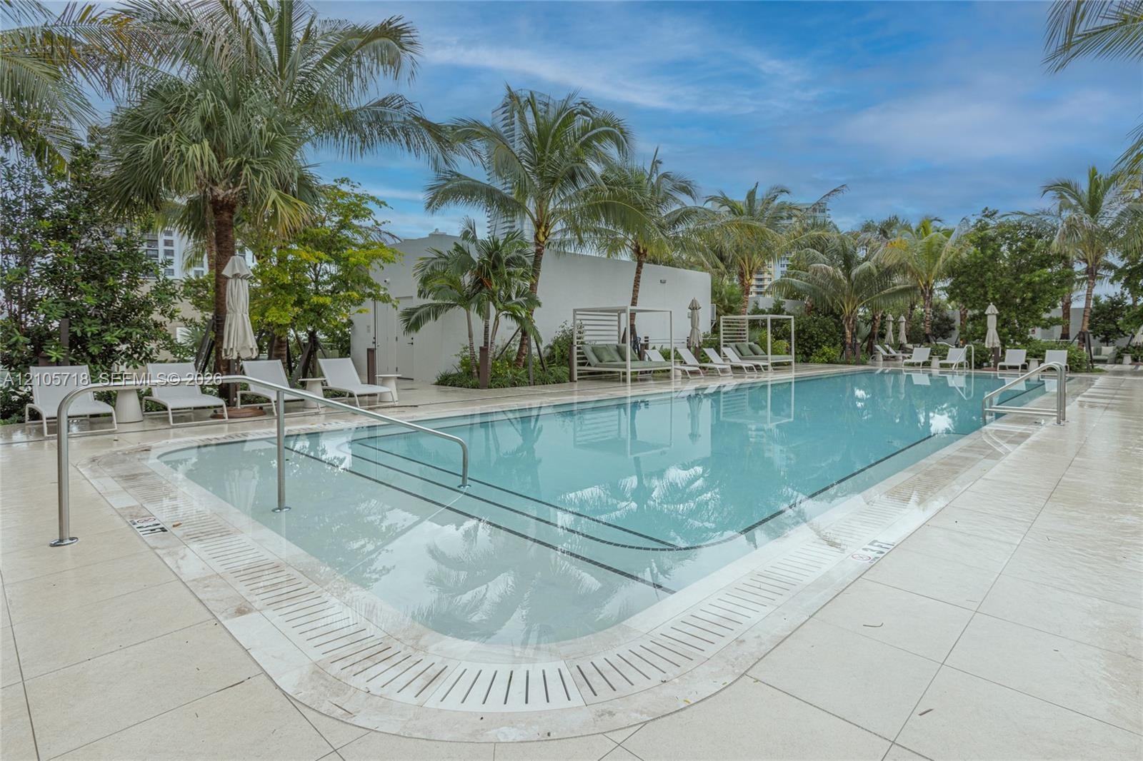 Rectangular swimming pool featuring a light-toned stone deck, integrated tiered steps, a stainless steel handrail, and mature palm tree landscaping