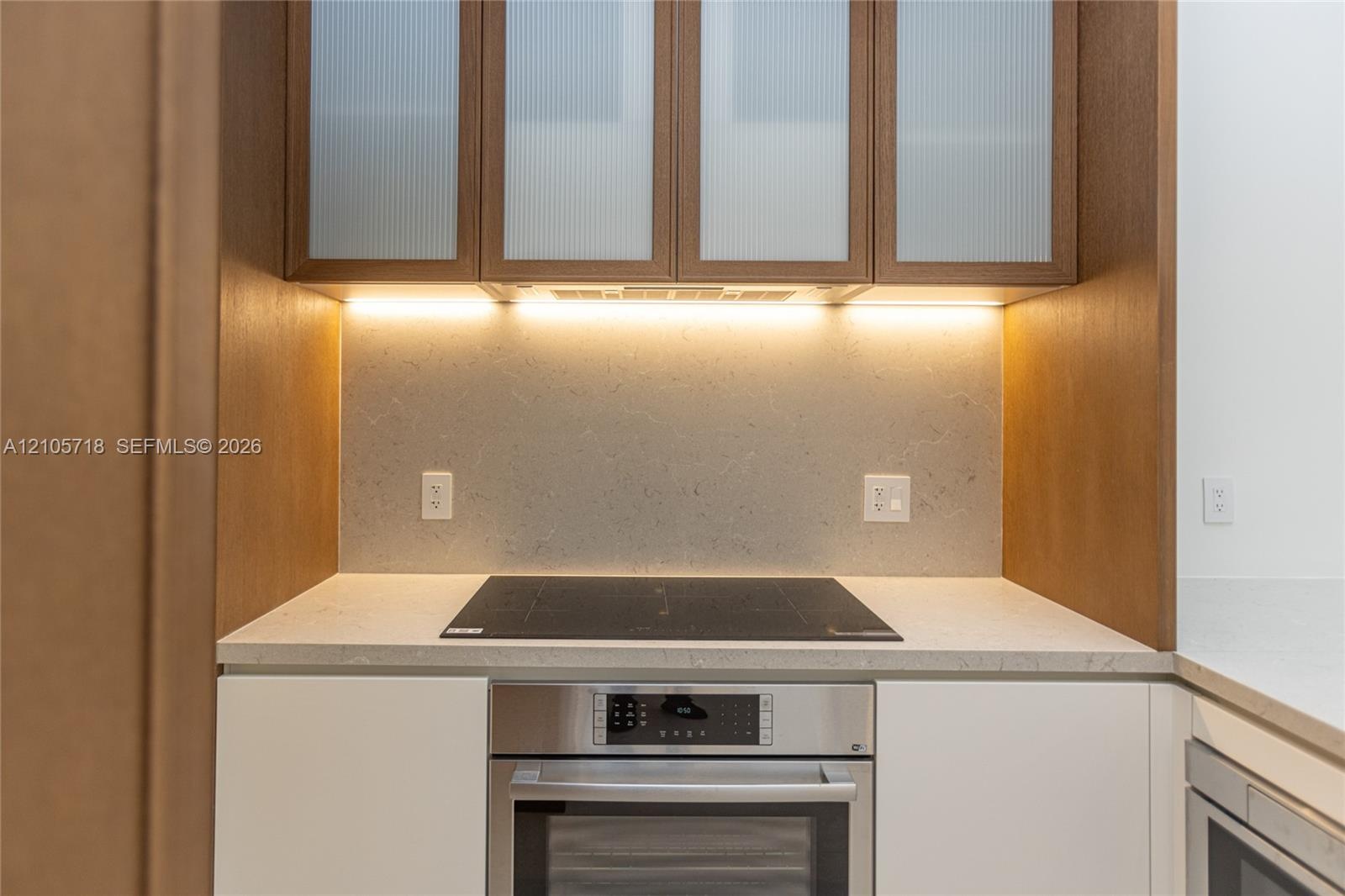 Kitchen area featuring matte white base cabinetry, integrated stainless steel oven, cooktop, solid-surface countertop, and wood-finish upper cabinets 