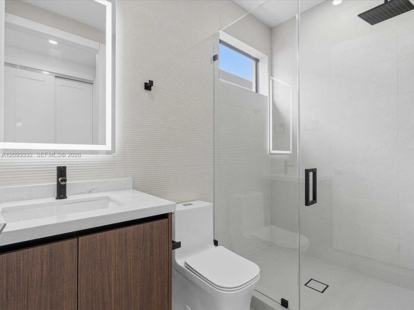 Modern vanity featuring a stone countertop, integrated sink, and illuminated mirror, paired with a glass-enclosed shower with matte black rainfall sho