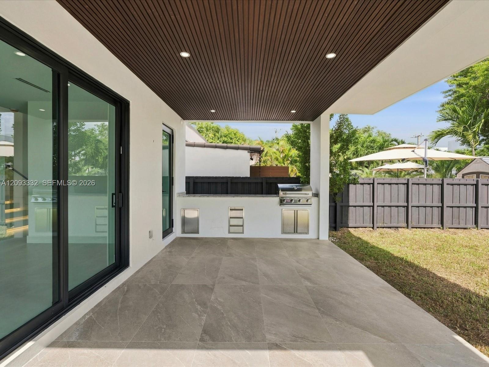 Covered patio featuring large-format stone-finish tile flooring, slatted wood-tone ceiling, recessed lighting, and a built-in outdoor kitchen with a s