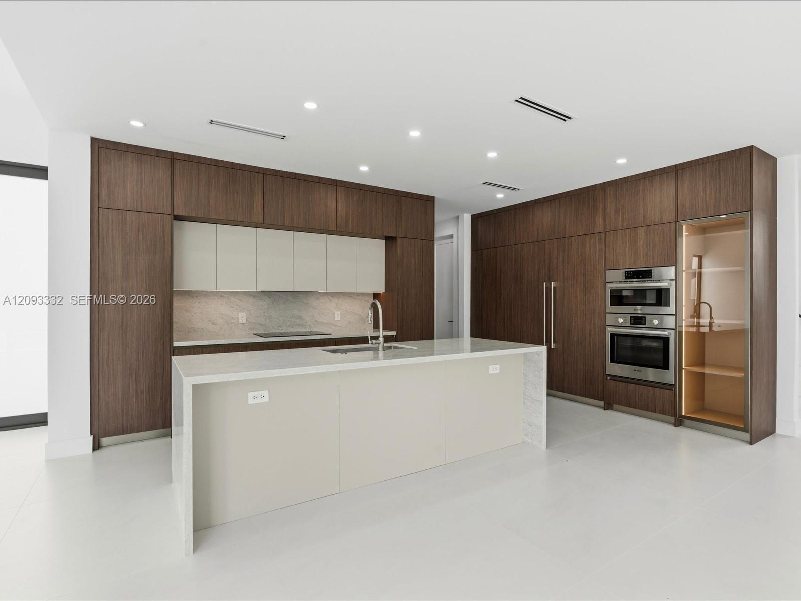 Modern kitchen featuring full-height wood-finish cabinetry, a waterfall-edge stone island, built-in stainless steel double ovens, and a glass-front di