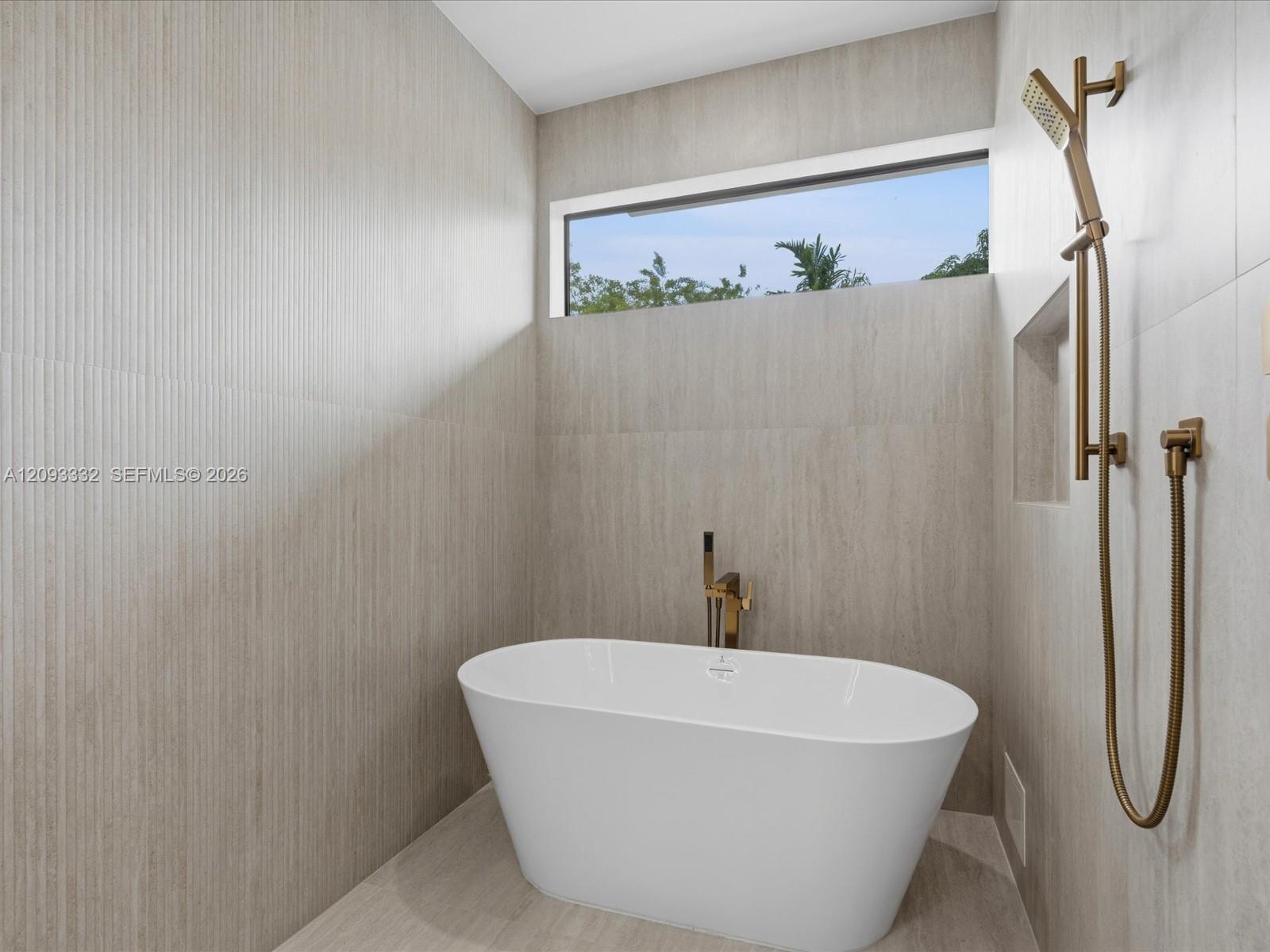Freestanding soaking tub featuring a metallic bronze floor-mounted faucet, textured stone-finish wall tile, a recessed wall niche, and a metallic bron