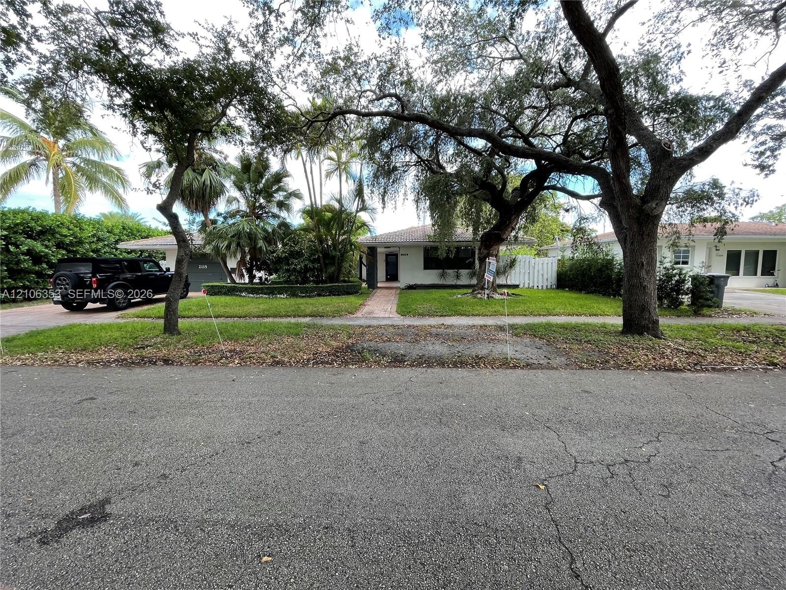 Single-story residence featuring a brick-paved walkway, mature canopy trees, manicured hedges, and an attached garage