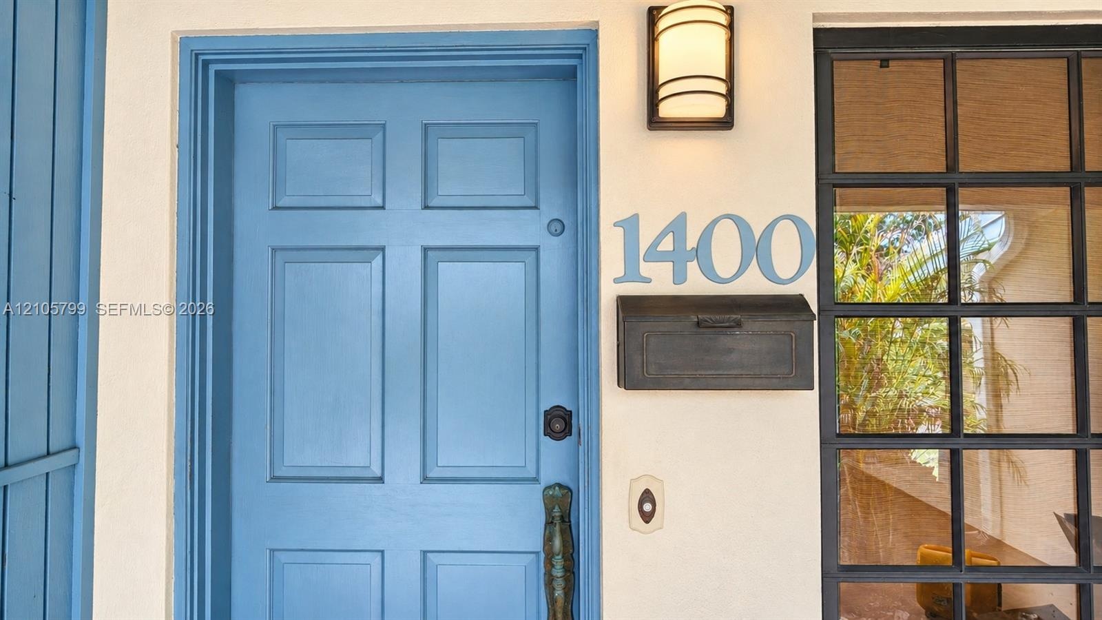 Entryway featuring a blue-painted paneled door, black metal wall-mounted mailbox, brushed metal house numbers, and a dark-framed grid-style sidelight