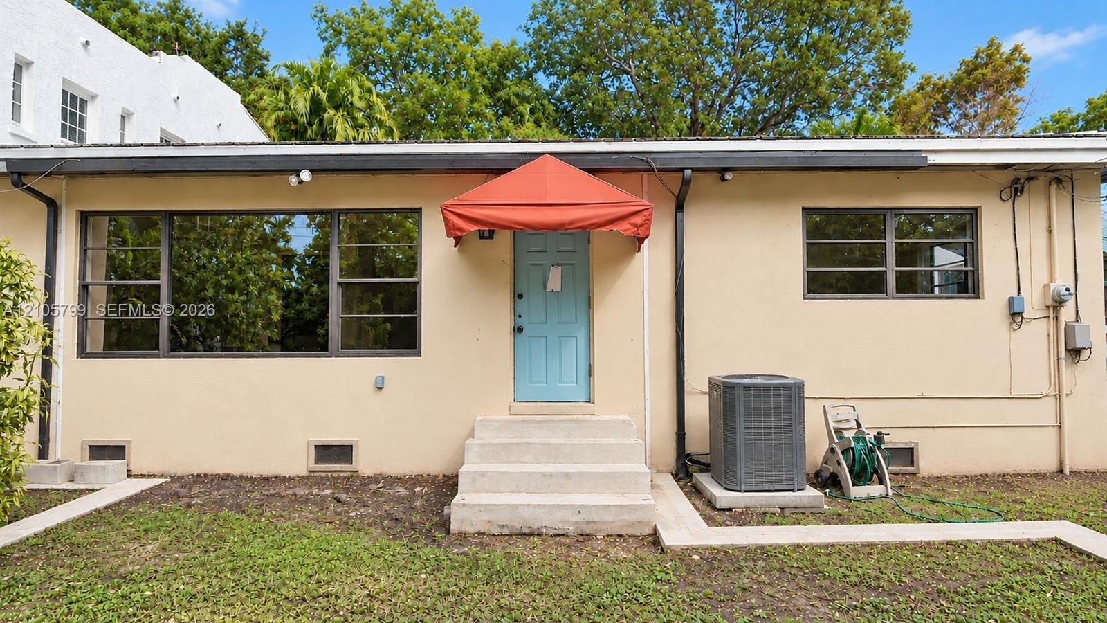 Stucco exterior featuring horizontal casement windows, a central entry with a fabric awning, concrete step access, and a dedicated outdoor heating and