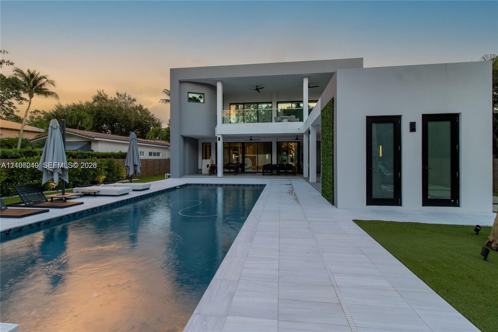 Modern two-story exterior featuring a rectangular in-ground pool, expansive light-colored stone patio, covered balcony with glass railing, and vertica