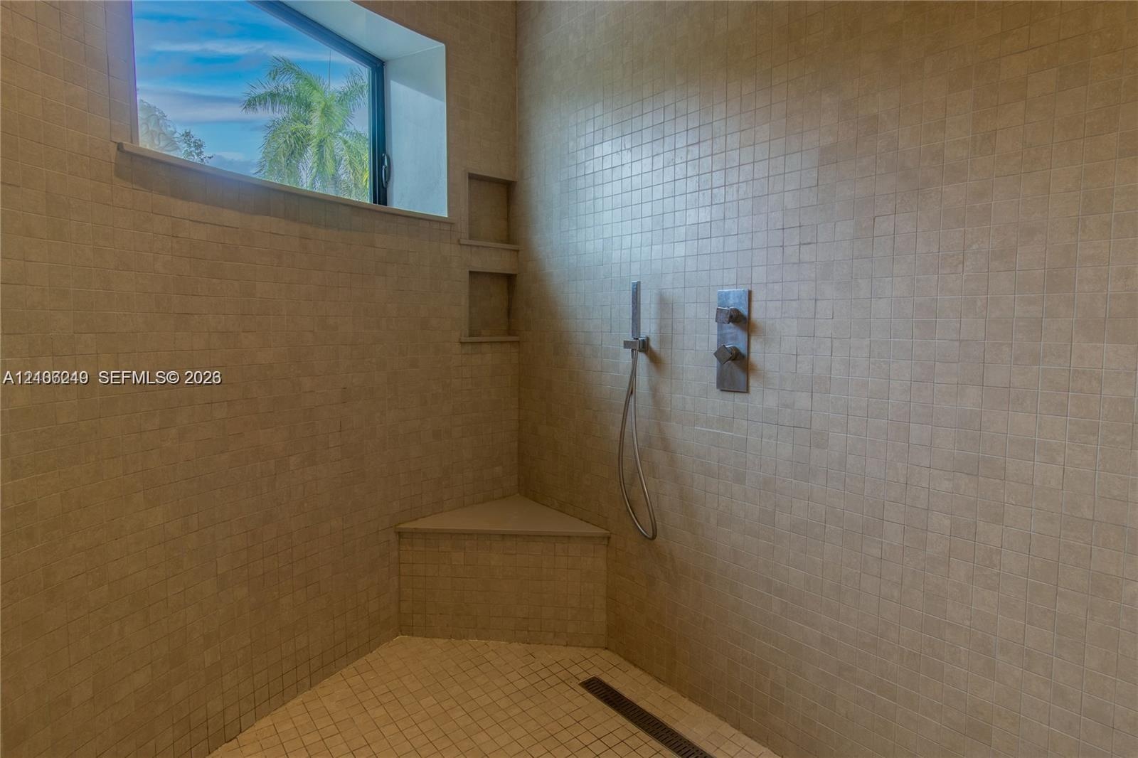 Walk-in shower featuring floor-to-ceiling mosaic tile, built-in corner bench, dual-recessed wall shelving, linear floor drain, and wall-mounted hand-h