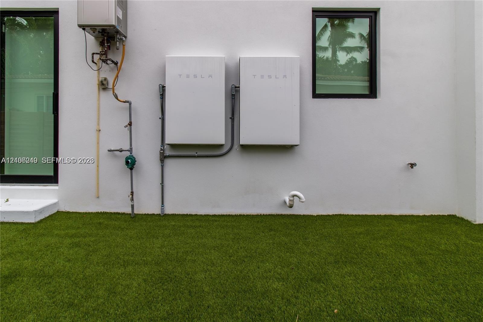 Exterior wall featuring dual Tesla Powerwall energy storage units, an on-demand tankless water heater, and artificial turf surfacing