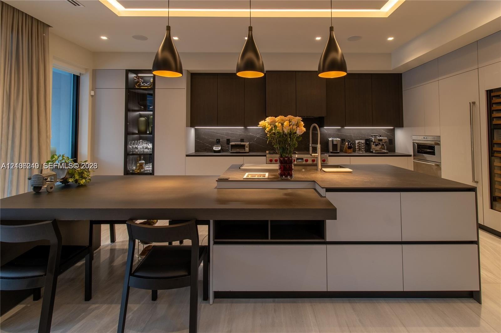 Modern kitchen featuring dark-toned cabinetry, a substantial kitchen island with an integrated dining surface, recessed ceiling lighting, three suspen