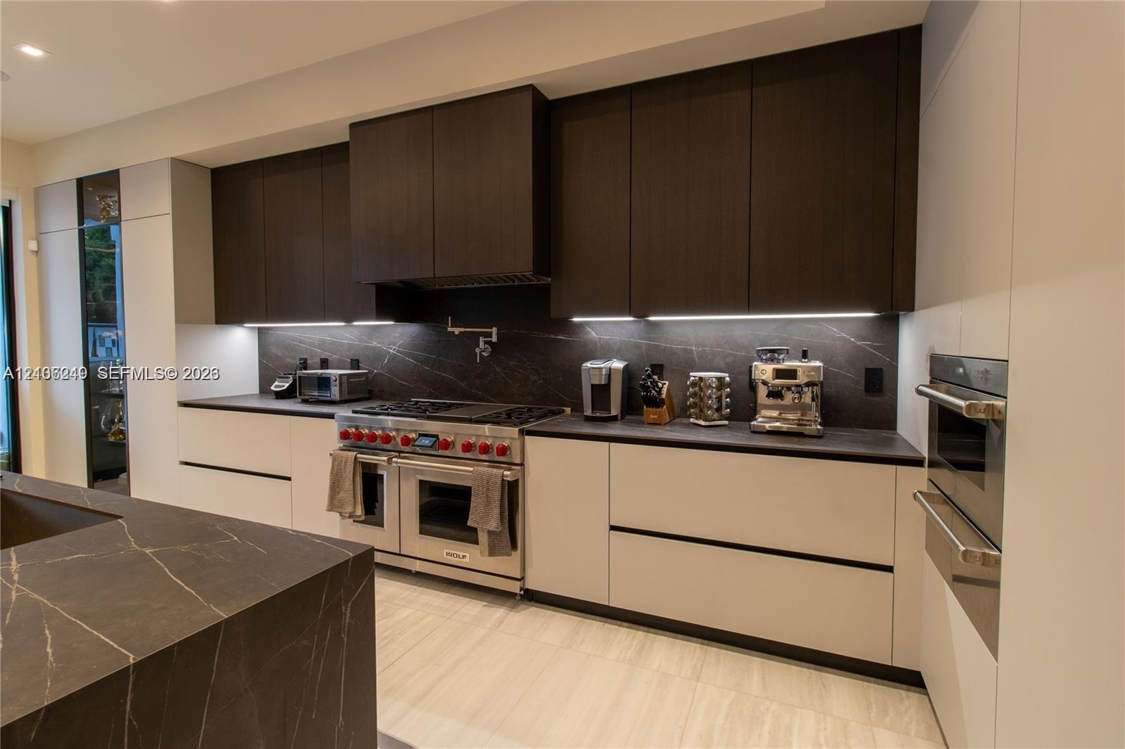 Modern kitchen featuring two-tone custom cabinetry, dark stone-finish countertops and backsplash, stainless steel gas range with pot filler, and built