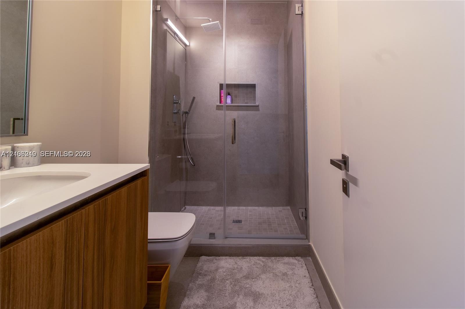 Wood-finish vanity cabinet with a white countertop and integrated sink, glass-enclosed shower featuring tiled walls, built-in recessed shelving, a rai