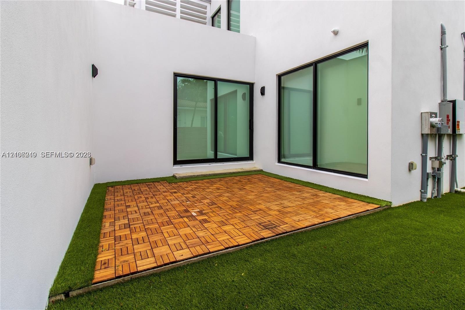 Modern outdoor patio featuring modular wood-finish decking, synthetic turf, minimalist white stucco exterior walls, and floor-to-ceiling sliding glass